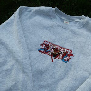May include: Grey sweatshirt with an embroidered image of three Spider-Man characters in red and blue, swinging from a web.