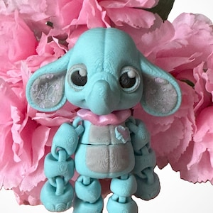 Puede incluir: Una figura de elefante articulada azul claro con orejas grandes y ojos grandes, que lleva una bufanda rosa y un chaleco gris con un corazón rosa. La figura se encuentra sobre un fondo de flores rosas.