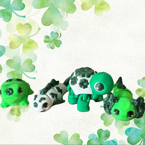 Könnte beinhalten: Drei grüne und weiße Figuren, die einen Frosch, ein Eidechse und eine Schildkröte darstellen. Die Figuren sind alle klein und im Cartoon-Stil gehalten. Sie sind alle mit Kleeblättern verziert.