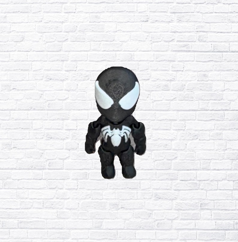 May include: A black and white figurine of a superhero, with a spider emblem on the chest. The figure has large white eye shapes and a spider symbol on the chest. The figurine is standing against a white brick wall.