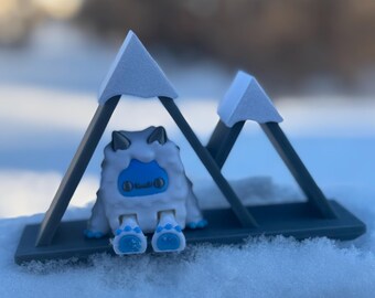 May include: A white and blue fuzzy toy with a blue face and blue feet sits on a gray mountain-shaped stand. The stand is covered in white snow.