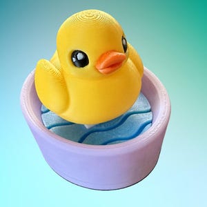 May include: A yellow rubber duck with big eyes and a small orange beak is floating in a light purple tub with blue wavy lines on the bottom.