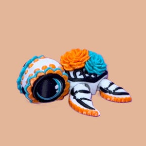 May include: A decorative turtle figurine with a white body, black accents, and orange and turquoise details. The turtle's shell is adorned with orange and turquoise floral designs. The figurine is set against a peach-colored background.