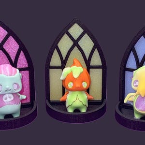May include: Three whimsical figurines displayed in arched window-shaped stands. The figurines are in the shape of cartoon characters with unique designs and colors. The windows have pink, yellow, and blue stained glass effects. The stands are dark purple.