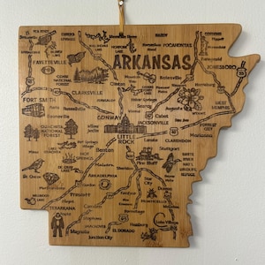May include: A wooden cutting board shaped like the state of Arkansas with the state's cities and towns engraved on it. The board is light brown with a natural wood grain.
