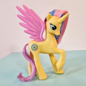 Könnte beinhalten: Eine gelbe My Little Pony Figur mit rosa Flügeln, einem blauen und gelben Symbol auf der Flanke und einer langen, rosa und blauen Mähne und einem Schwanz.