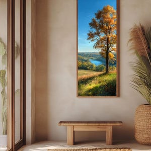 Puede incluir: Una pintura de paisaje vertical enmarcada representa un árbol con hojas doradas contra un cielo azul. La obra de arte muestra una vista panorámica de un río, campos y árboles distantes. La pintura se exhibe sobre un banco de madera y una alfombra tejida.
