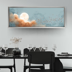 May include: A framed canvas art piece depicting a full moon, clouds, and birds in flight. The artwork features a silver frame and a color palette of blue, orange, and white. The artwork is displayed above a dining table with wine glasses.