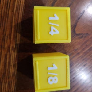 May include: Two yellow plastic measuring cups with white numbers. The top cup says "1/4" and the bottom cup says "1/8".
