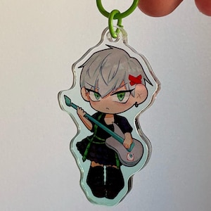 May include: A clear acrylic keychain featuring a cartoon character with silver hair, green eyes, and a red bow. The character is wearing a black dress and holding a green and white guitar.