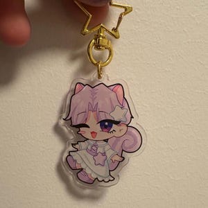 May include: A purple and white anime character keychain with a star on its head. The character is wearing a white dress with a star on it. The keychain is attached to a gold star-shaped charm.