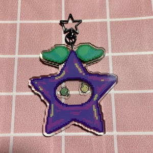 May include: A purple star-shaped keychain with two green eyes and a green plant on top. The keychain is made of clear acrylic and has a pixelated design.