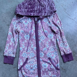 K&ouml;nnte beinhalten: Ein lila-wei&szlig; gemusterter Kapuzenpullover mit Rei&szlig;verschluss. Der Hoodie hat lange &Auml;rmel und zwei Taschen.