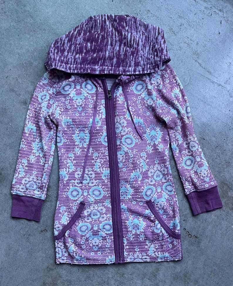 K&ouml;nnte beinhalten: Ein lila-wei&szlig; gemusterter Kapuzenpullover mit Rei&szlig;verschluss. Der Hoodie hat lange &Auml;rmel und zwei Taschen.