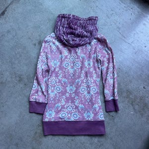 K&ouml;nnte beinhalten: Ein lang&auml;rmliger Kapuzenpullover mit einem Muster in Wei&szlig; und Lila. Die Kapuze ist mit einem Tie-Dye-Muster in Wei&szlig; und Lila gef&uuml;ttert.