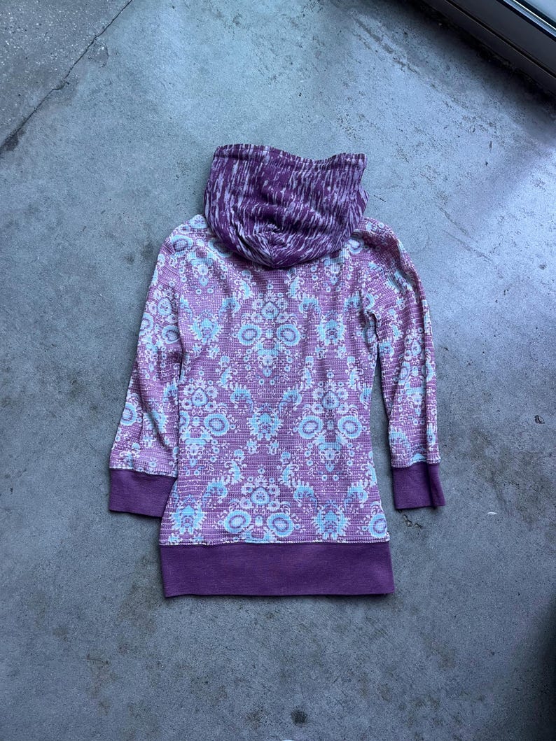 K&ouml;nnte beinhalten: Ein lang&auml;rmliger Kapuzenpullover mit einem Muster in Wei&szlig; und Lila. Die Kapuze ist mit einem Tie-Dye-Muster in Wei&szlig; und Lila gef&uuml;ttert.