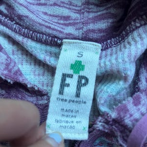 K&ouml;nnte beinhalten: Ein lila-wei&szlig; gestreiftes Kapuzenpullover mit einem Free People-Etikett. Das Etikett tr&auml;gt die Aufschrift "Free People" und "Hergestellt in Macau" in schwarzer Schrift. Das Etikett hat auch ein gr&uuml;nes Pluszeichen und den Buchstaben "S" f&uuml;r die Gr&ouml;&szlig;e.