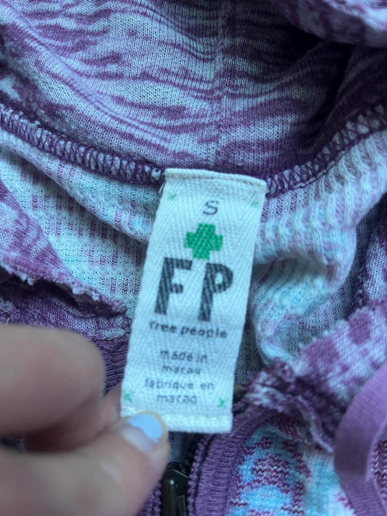 K&ouml;nnte beinhalten: Ein lila-wei&szlig; gestreiftes Kapuzenpullover mit einem Free People-Etikett. Das Etikett tr&auml;gt die Aufschrift "Free People" und "Hergestellt in Macau" in schwarzer Schrift. Das Etikett hat auch ein gr&uuml;nes Pluszeichen und den Buchstaben "S" f&uuml;r die Gr&ouml;&szlig;e.