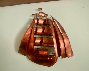 Pode incluir: Arte de parede em metal cor de cobre representando um veleiro. O navio apresenta velas detalhadas, um casco com vigias e um mastro com bandeiras. A obra de arte é montada em uma parede de cor clara, criando um contraste marcante. Esta decoração com tema náutico adiciona charme marítimo.