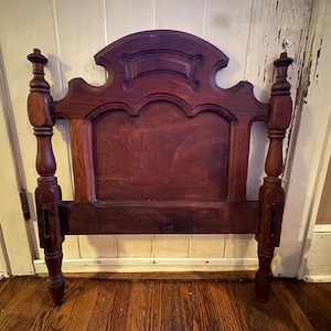 Puede incluir: Un cabecero de madera marrón oscuro con un panel central rectangular y una cresta superior curva. El marco incluye dos postes verticales con detalles torneados y remates decorativos en la parte superior, junto con ranuras metálicas.