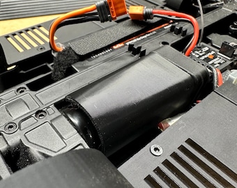 May include: Close-up of a black RC car chassis with an orange and red wire connected to a battery pack. The battery pack is secured in a black plastic housing. The chassis has a black plastic cover with a vent.