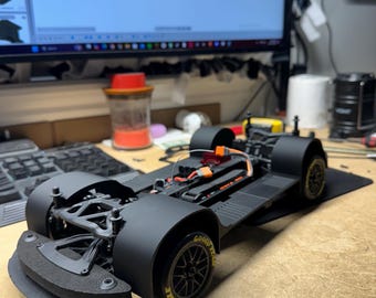 May include: A black and gray race car chassis with black wheels and Goodyear Eagle tires. The chassis is on a wooden surface.