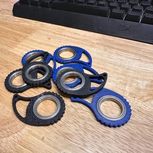 May include: A collection of black and blue fidget spinners with a circular design and a silver bearing. The spinners are made of plastic and have a textured surface. The background includes a wooden surface and a black keyboard.