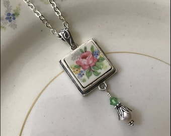 Peut inclure: Un collier en chaîne argent avec un pendentif carré orné d'un motif floral vintage. Le pendentif présente une rose rose et des feuilles vertes sur fond blanc. Un seul cristal vert et une perle blanche pendent du bas du pendentif.