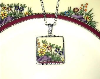 Könnte beinhalten: Eine quadratische Anhängerhalskette mit floralem Design. Der Anhänger hat einen cremefarbenen Hintergrund mit gelben, blauen und orangen Blumen, grünen Blättern und lila Felsen. Der Anhänger ist silberfarben gerahmt und hängt an einer silbernen Kette.
