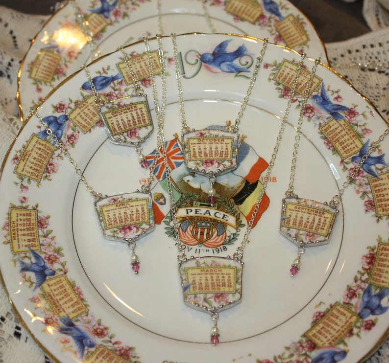 K&ouml;nnte beinhalten: Eine Sammlung von Vintage-Kalenderketten mit silbernen Ketten und Anh&auml;ngern, die Monatskalender von April bis September zeigen. Die Anh&auml;nger sind mit Blumendesigns verziert und haben eine Vintage-&Auml;sthetik. Die Ketten werden auf einem wei&szlig;en Porzellanteller mit Blumenmuster pr&auml;sentiert.