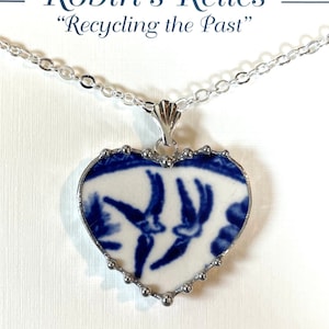 May include: A silver chain necklace with a heart-shaped pendant. The pendant is made from a vintage blue and white china plate with a design of two birds in flight. The pendant is surrounded by a silver beaded border.