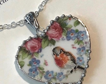 Peut inclure: Un pendentif en forme de cœur représentant un rouge-gorge entouré de roses roses et de fleurs bleues sur fond blanc. Le pendentif est enchâssé dans un cadre en métal argenté avec un bord perlé et est suspendu à une chaîne en argent.