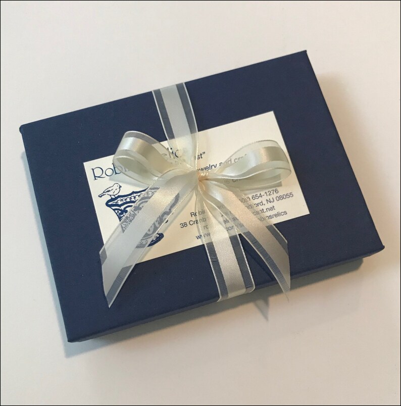 K&ouml;nnte beinhalten: Eine dunkelblaue Geschenkbox mit einem wei&szlig;en Satinband und Schleife. Die Box hat ein wei&szlig;es Etikett mit dem Text "Rob's Relics" und Kontaktinformationen.
