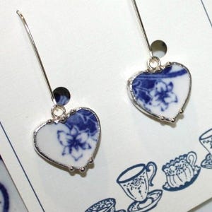 Puede incluir: Pendientes en forma de corazón con un diseño floral azul sobre fondo blanco, enmarcados en plata. Los pendientes cuelgan de ganchos plateados. El diseño recuerda a la porcelana vintage. El fondo presenta ilustraciones de tazas de té.