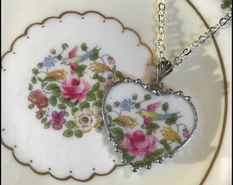 May include: Heart-shaped pendant necklace with a floral design featuring pink roses, blue and yellow birds, and green leaves. The pendant is framed in silver and hangs on a silver chain. The necklace rests on a floral patterned plate.