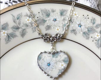 Peut inclure: Un collier argenté avec un pendentif en forme de cœur orné d'un motif floral crème et d'un accent bleu au centre. Le pendentif est fixé à une chaîne argentée par une pièce métallique décorative représentant de petites fleurs et feuilles.