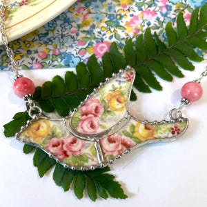 May include: A handcrafted necklace featuring a pendant made from a vintage floral china plate. The pendant showcases pink and yellow roses, framed in silver. The necklace includes pink gemstone beads and a delicate silver chain.