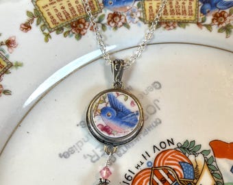 Könnte beinhalten: Halskette mit silberfarbenem Anhänger mit einer Illustration eines blauen Vogels. Eine kleine rosa Perle und ein perlenartiger Anhänger hängen darunter. Die Kette liegt auf einem Vintage-Teller mit Blumen- und Vogelmotiven.