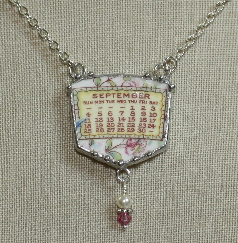 K&ouml;nnte beinhalten: Eine Halskette aus Silberkette mit einem Anh&auml;nger im Vintage-Kalender-Design. Der Anh&auml;nger ist aus wei&szlig;em Porzellan mit einem Blumenmuster und einem roten und wei&szlig;en Kalender f&uuml;r den Monat September. Der Anh&auml;nger ist an einer Silberkette mit einer Perle und einem rosafarbenen Kristall befestigt.