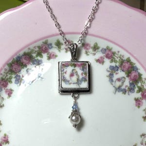 May include: A silver chain necklace featuring a square pendant with a white ceramic tile insert depicting a bird surrounded by pink roses. A small silver bead cap holds a white pearl and a tiny blue crystal bead dangling from the bottom of the pendant.