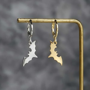 May include: A pair of hoop earrings featuring bat-shaped charms. One earring is silver-toned and the other is gold-toned. Each charm is suspended from a small circular hoop, showcasing a detailed silhouette of a bat with wings spread wide.