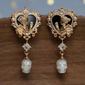 May include: Gold earrings with a black heart design featuring a cherub and a dangling pearl skull charm.