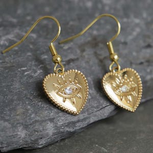 May include: Gold heart-shaped earrings with a small, clear crystal eye in the center. The earrings have a delicate, detailed design and are perfect for adding a touch of sparkle to any outfit.