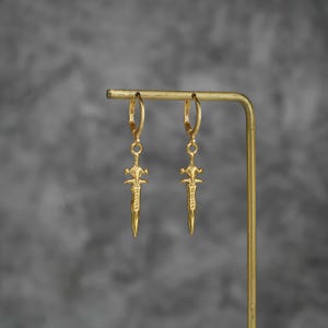 May include: A pair of gold-toned hoop earrings featuring dangling sword-shaped charms. Each earring consists of a circular hoop connected to a detailed, pointed blade pendant with a cross-guard design, suspended from a thin metal display stand.