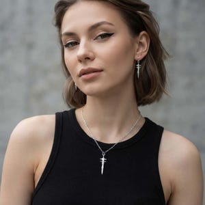 May include: A silver-toned metal necklace featuring a sword-shaped pendant on a delicate chain, paired with matching sword-shaped drop earrings. The jewellery set is worn against a plain black sleeveless top, showcasing a polished metallic finish.