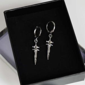 May include: A pair of silver-toned hoop earrings featuring dangling dagger-shaped charms. Each charm has a pointed blade design with a cross-guard and a textured handle. The earrings are displayed inside a black box with a velvet-like interior lining.
