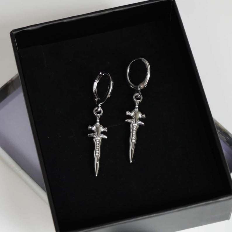 May include: A pair of silver-toned hoop earrings featuring dangling dagger-shaped charms. Each charm has a pointed blade design with a cross-guard and a textured handle. The earrings are displayed inside a black box with a velvet-like interior lining.