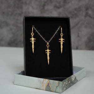 May include: A gold-toned jewellery set consisting of a necklace and two matching earrings, each featuring a small sword-shaped pendant. The pieces are displayed inside a black rectangular box with a light-coloured, marbled-patterned base.