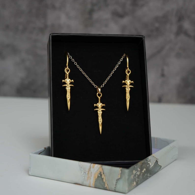 May include: A gold-toned jewellery set consisting of a necklace and two matching earrings, each featuring a small sword-shaped pendant. The pieces are displayed inside a black rectangular box with a light-coloured, marbled-patterned base.
