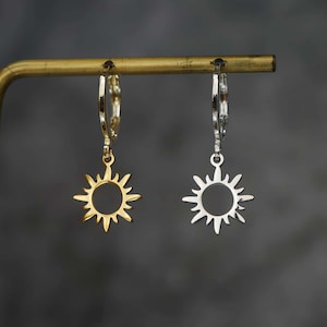 May include: Two sun-shaped earrings are shown, one gold and one silver. Each earring has a central circle with outward rays. They hang from a gold-coloured bar against a dark grey backdrop. The earrings are likely made of metal.
